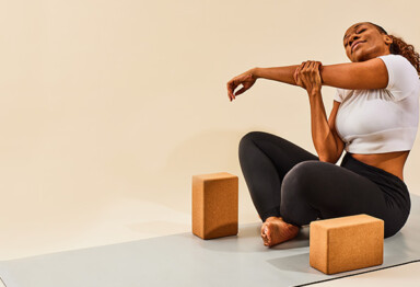 Woman sitting on yoga mat with yoga blocks doing arm stretches and smiling