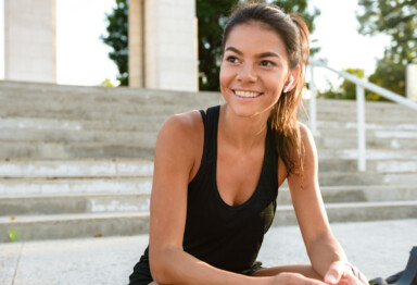 Woman smiling sitting on concrete steps outside mid workout wearing ear buds.